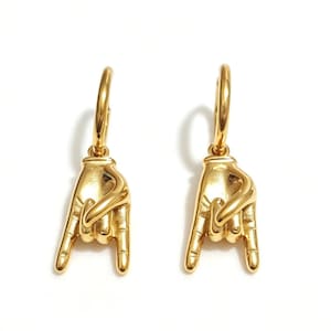 May include: A pair of gold-toned hoop earrings featuring a dangling charm shaped like a hand making a rock and roll gesture. The hand charm has detailed fingers and a smooth, polished metallic finish, suspended from a simple circular hoop.