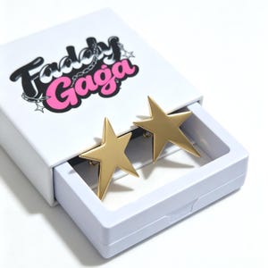 May include: A pair of gold-toned star-shaped earrings resting inside a white sliding box. The box features the text Faddy Gaga in black and pink lettering with a small chain graphic. The earrings have a smooth, matte finish and five points.