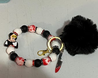 Könnte beinhalten: Ein Perlen-Schlüsselanhänger-Armband mit schwarzen, weißen und roten Perlen. Das Armband hat einen Anhänger einer Zeichentrickfigur, einen goldfarbenen Verschluss und einen kleinen Messeranhänger. Ein schwarzer, flauschiger Pompon ist befestigt.