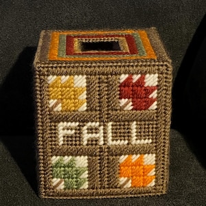 May include: A brown, square tissue box cover with the word "FALL" stitched in white. The cover features four squares with leaf designs in yellow, red, green, and orange. The top has a rectangular opening and is trimmed with various colors.