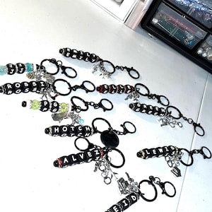 May include: A collection of black beaded keychains with white lettered names, including "EMILY", "ROSSI", and "HOTCH". Each keychain has a silver charm and a black keyring. The background includes storage containers filled with beads and glitter.