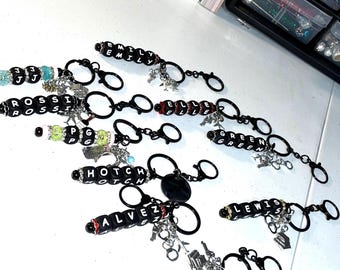 May include: A collection of black keychains featuring letter beads spelling names like EMILY, ROSSI, ELLE, GREEN, HOTCH, ALVEZ, LEWIS, and REID. Each keychain includes silver-toned charms, black rings, and decorative beads in various colors.