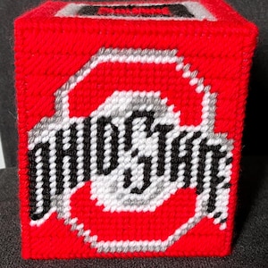 May include: A red, handmade tissue box cover featuring the Ohio State Buckeyes logo. The logo is in white, black, and grey, with the words "OHIO STATE" displayed. The box is square and made of a woven material.