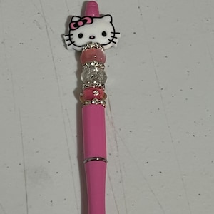 Könnte beinhalten: Ein pinkfarbener Stift mit einem Hello Kitty-Charakter oben. Der Stift hat rosa und klare Perlen. Der Schaft ist einfarbig rosa mit einem silbernen Band. Die Stiftspitze ist silberfarben.