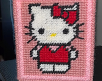 May include: A rectangular plastic canvas needlepoint project featuring a stitched Hello Kitty character. The figure has a white face, black eyes, a yellow nose, and a red bow, wearing a red top. The border is stitched in light pink yarn.