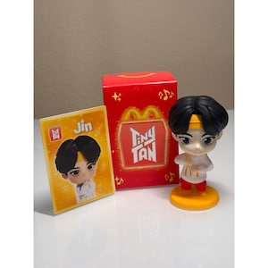 May include: A Tiny Tan figurine of Jin, with black hair and a yellow headband, stands on a yellow base. A red box with the Tiny Tan logo and a card with Jin's image are also present. The McDonald's logo is visible.