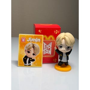 May include: A collectible set featuring a Tiny Tan Jimin figure, a card, and a red box. The figure has a blonde hairstyle, black jacket, and yellow base. The card and box feature the Tiny Tan and Jimin logos.