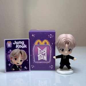 May include: A set of Tiny Tan collectibles featuring a cartoon figure, a card, and a purple box. The figure has light-colored hair and is wearing a black suit. The card and box feature the Tiny Tan logo and the name "Jungkook".