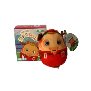 May include: A plush toy named Maple, a brown deer character with antlers, wearing a red soccer jersey with FIFA branding. Beside it is a cardboard box featuring the Original Squishmallows logo, the number 5, and an illustration of the same character.
