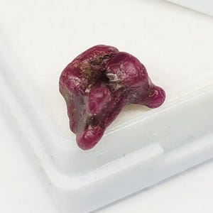 May include: A raw, unpolished ruby gemstone with a deep magenta and dark red coloration. The stone features an irregular, organic shape with multiple rounded protrusions and a textured surface showing natural mineral inclusions and variations.
