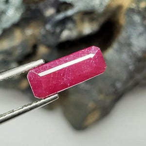 May include: A faceted, rectangular ruby gemstone held by silver tweezers. The ruby is a deep pinkish-red colour with a white, linear inclusion running along its length. The background is a blurred, dark grey and brown.