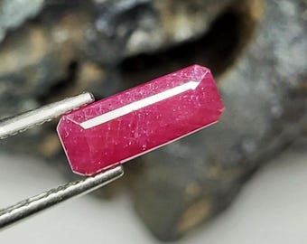 May include: A faceted, rectangular ruby gemstone held by silver tweezers. The ruby is a deep pinkish-red color with a white, linear inclusion running along its length. The background is a blurred, dark gray and brown.