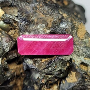 May include: A vibrant, rectangular-cut ruby gemstone. The gem is a deep, saturated pink-red color, with visible internal facets. It rests on a rough, textured, dark-colored rock surface, highlighting the gem's brilliance.