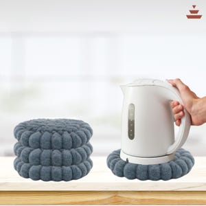 May include: Three stacked, round, gray felt trivets next to a white electric kettle. The trivets have a textured, raised design. The kettle is on a matching trivet. The background is a light, blurred interior.