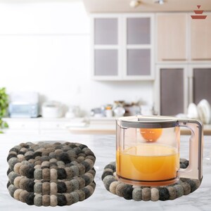 Puede incluir: Juego de posavasos de fieltro en tonos grises, marrones y negros. Una jarra transparente con líquido naranja descansa sobre un posavasos a juego. Escena de cocina con armarios blancos y encimera.