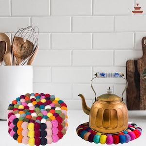 May include: Two felt ball trivets are shown. One is a round trivet with a variety of colours, including pink, yellow, green, and blue. The other trivet is under a brass tea kettle. Kitchen decor.