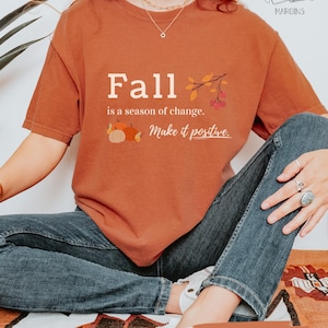 May include: A burnt orange t-shirt with the text "Fall is a season of change. Make it positive." and a fall-themed graphic. The shirt is paired with blue jeans. The shirt is labeled "YAM".