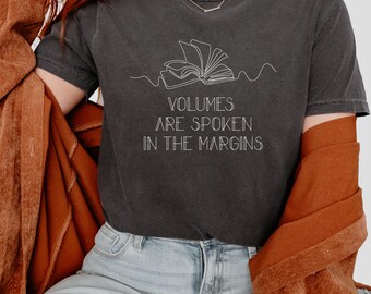 May include: Dark gray t-shirt with the text "VOLUMES ARE SPOKEN IN THE MARGINS" in white. The shirt features a line drawing of an open book. The model is wearing blue jeans and a brown jacket.