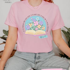 May include: A light pink t-shirt with a floral design featuring an open book and the text "Let us therefore make every effort to do what leads to peace and to mutual edification." The text "Romans 14:19" is also included.