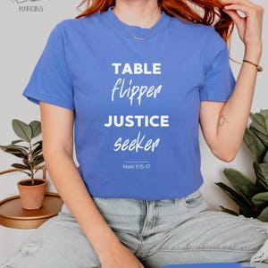 Puede incluir: Una camiseta azul con las palabras "TABLE FLIPPER JUSTICE seeker" en blanco, con "Mark 11:15-17" debajo. La camiseta tiene cuello redondo y mangas cortas. El texto "FLO BLUE" está escrito en una franja azul.