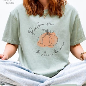 以下が含まれることがあります： 「Pumpkin spice & please be nice」の文字とカボチャの線画が描かれたセージグリーンのTシャツ。半袖でゆったりとしたフィット感。ブランド名「Volumes in the Margins」が左上にあります。