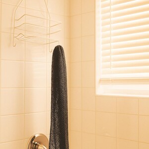 May include: A black mesh shower sponge hanging in a tiled bathroom. The sponge is suspended from a shower fixture. A white wire shower caddy with shelves is mounted above the sponge. A window with blinds is visible.