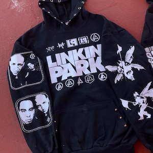 Puede incluir: Sudadera negra con el nombre de la banda "LINKIN PARK" en letras blancas. Las mangas muestran retratos de los miembros de la banda y diseños gráficos. La capucha tiene ojales metálicos. Prenda cómoda para fans.