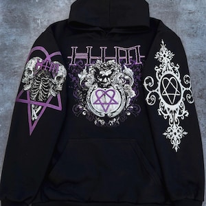 May include: A black hooded sweatshirt featuring purple and white graphic prints. The front displays a central ornate emblem with a heart-shaped pentagram and text. The sleeves feature a skull design on the left and a decorative cross on the right.