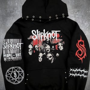 Może przedstawiać: Czarna bluza z kapturem z dużą grafiką zespołu Slipknot na piersi. Na rękawach znajduje się kod kreskowy z napisem PEOPLE=SHIT, symbol nonagramu, czerwone logo S w stylu plemiennym oraz grafika z drutem kolczastym. Srebrne oczka przy kapturze.