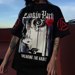 Può includere: Una t-shirt nera a maniche corte con una stampa grafica di un volto stilizzato e il testo Linkin Park e Breaking The Habit. Le maniche presentano un logo circolare rosso. Il design include inchiostro nero, bianco e rosso su base nera.