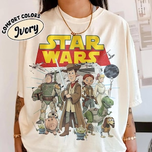 Puede incluir: Camiseta de color marfil con un gr&aacute;fico de personajes de Star Wars de estilo caricatura. El logotipo de Star Wars es amarillo y rojo. La camiseta tambi&eacute;n presenta las palabras "Comfort Colors" e "Ivory" en un dise&ntilde;o circular.