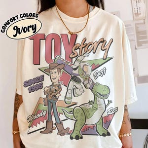 May include: Ivory t-shirt with the words "Toy Story" in maroon and gold, featuring Woody, Buzz, and Rex. The design includes "Since 1995" and "Comfort Colors" in a circular design. The shirt is a cream color.