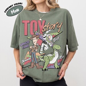 May include: Moss green t-shirt with the Toy Story logo and characters Woody, Buzz, and Rex. The design includes the text "TOY STORY" and "SINCE 1995". The shirt has a vintage, distressed appearance.