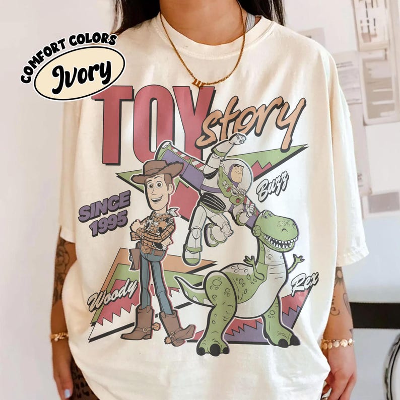 May include: Ivory t-shirt with the words "Toy Story" in maroon and gold, featuring Woody, Buzz, and Rex. The design includes "Since 1995" and "Comfort Colors" in a circular design. The shirt is a cream color.