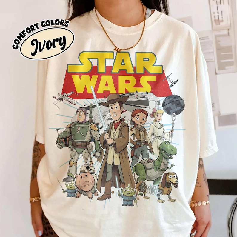 Puede incluir: Camiseta de color marfil con un gr&aacute;fico de personajes de Star Wars de estilo caricatura. El logotipo de Star Wars es amarillo y rojo. La camiseta tambi&eacute;n presenta las palabras "Comfort Colors" e "Ivory" en un dise&ntilde;o circular.