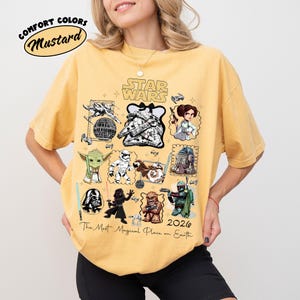 K&ouml;nnte beinhalten: Senffarbenes T-Shirt mit einem Star Wars-Motiv, das verschiedene Charaktere und den Text "The Most Magical Place on Earth 2026" zeigt. Das Shirt hat auch die W&ouml;rter "Comfort Colors" und "Mustard" in einem Kreis.