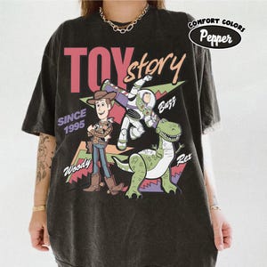 May include: Black t-shirt with a Toy Story design, showcasing Woody, Buzz Lightyear, and Rex. The graphic includes the words "TOY STORY" in pink and white, "SINCE 1995", and character names. The shirt is from Comfort Colors.