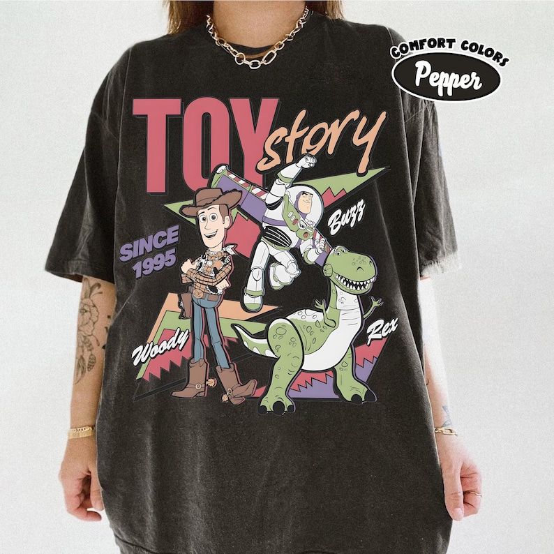 May include: Black t-shirt with a Toy Story design, showcasing Woody, Buzz Lightyear, and Rex. The graphic includes the words "TOY STORY" in pink and white, "SINCE 1995", and character names. The shirt is from Comfort Colors.