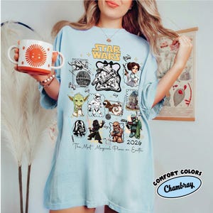 K&ouml;nnte beinhalten: Hellblaues T-Shirt mit Star Wars-Charakteren und dem Text "Star Wars 2026 The Most Magical Place on Earth." Das Shirt hat auch das Comfort Colors Chambray-Logo und eine Tasse mit einem Sonnendesign.