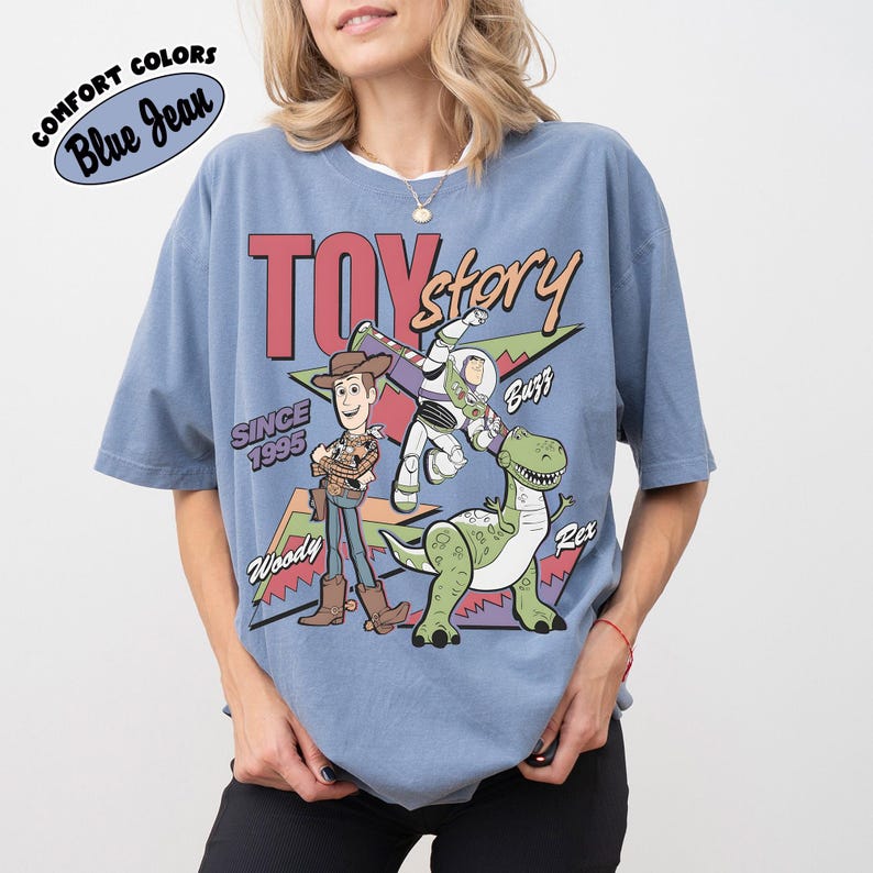 May include: Blue short-sleeved t-shirt with the Toy Story logo and Woody, Buzz Lightyear, and Rex characters. The shirt includes the text "Since 1995" and "Comfort Colors Blue Jean" in a circular design.