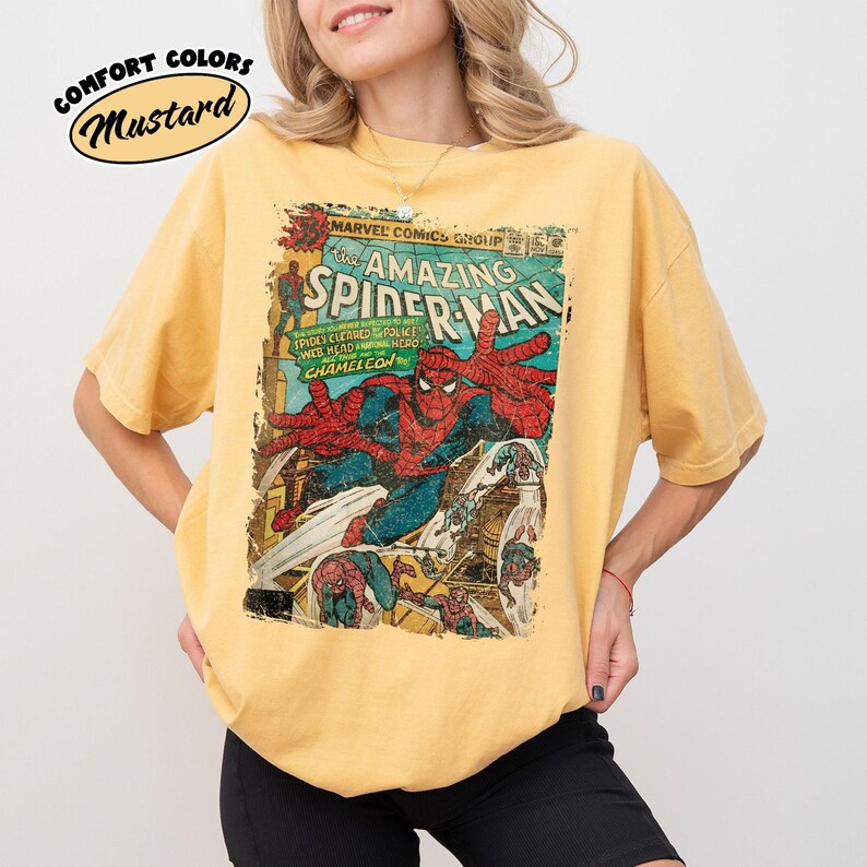 May include: Mustard-colored t-shirt featuring a vintage comic book cover of The Amazing Spider-Man. The graphic includes Spider-Man in action, with text that reads "Marvel Comics Group" and "Spidey Cleared Police Web Head Hero Chameleon."