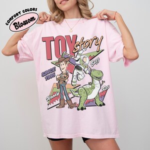 May include: Pale pink t-shirt featuring Woody, Buzz Lightyear, and Rex from Toy Story. The design includes the words "TOY STORY" and "SINCE 1995". The shirt is labelled "Comfort Colors Blossom".