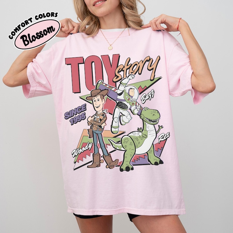 May include: Pale pink t-shirt featuring Woody, Buzz Lightyear, and Rex from Toy Story. The design includes the words "TOY STORY" and "SINCE 1995". The shirt is labelled "Comfort Colors Blossom".