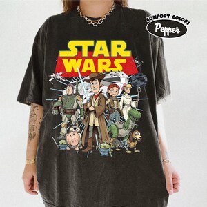 Puede incluir: Camiseta gris oscuro con un gr&aacute;fico de personajes de Star Wars, incluyendo a Woody y Buzz Lightyear. El logo de Star Wars es amarillo y rojo. La camiseta est&aacute; etiquetada como "Comfort Colors Pepper".