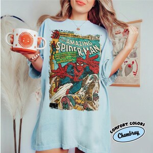May include: A light blue t-shirt featuring a vintage comic book cover of Spider-Man. The shirt has the text "AMAZING SPIDER-MAN" and "MARVEL COMICS GROUP". The shirt is labeled "COMFORT COLORS Chambray". A white mug with an orange sun design is held in the image.