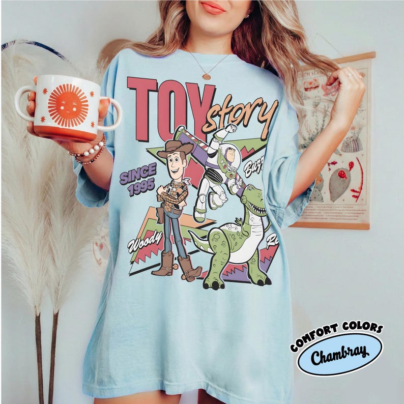 May include: Pale blue t-shirt with a "Toy Story" design, including Woody, Buzz Lightyear, and Rex characters. The shirt also features the text "Since 1995" and "Comfort Colors Chambray" printed on it.