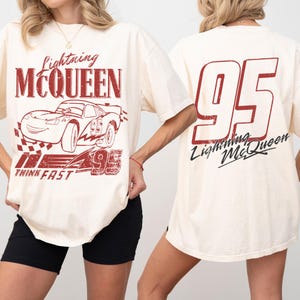 May include: Cream-coloured t-shirt with a distressed graphic of Lightning McQueen race car, the text "Lightning McQueen" and "95". The back of the shirt displays a large "95" and "Lightning McQueen" text.