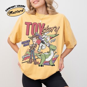 May include: Mustard-coloured t-shirt featuring the Toy Story logo and characters Woody, Buzz Lightyear, and Rex. The shirt has the text "Since 1995" and "Comfort Colors" in a circular design.