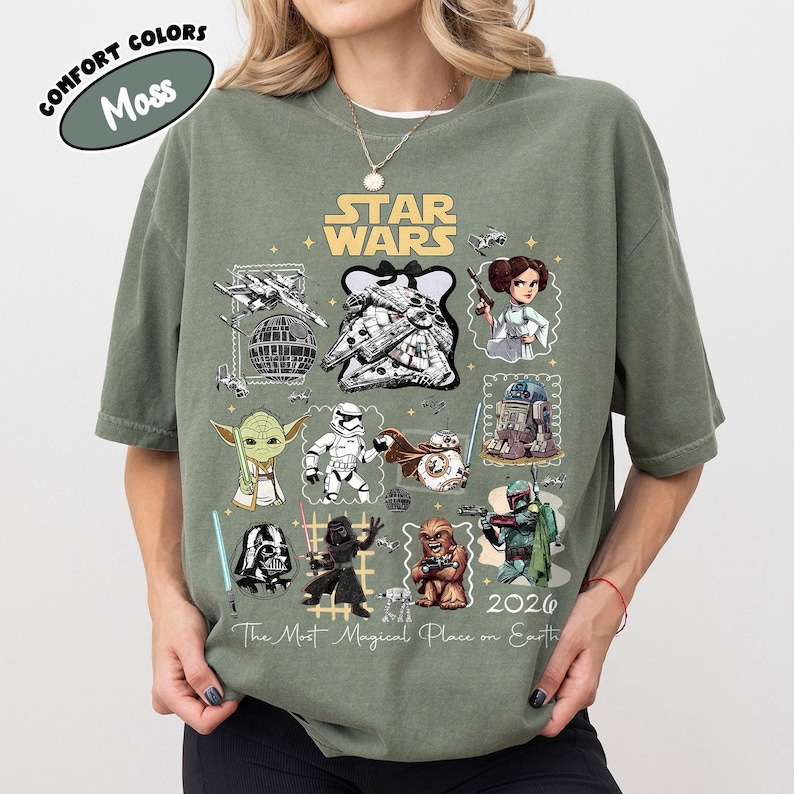 K&ouml;nnte beinhalten: Moosgr&uuml;nes Star Wars T-Shirt mit verschiedenen Charakteren und Elementen aus dem Franchise, darunter Yoda, Darth Vader und der Millennium Falke. Das Shirt hat den Text "Star Wars" und "2026 The Most Magical Place on Earth."