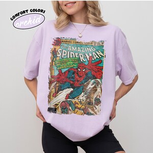 May include: A light purple t-shirt featuring a vintage comic book cover of The Amazing Spider-Man. The comic book art shows Spider-Man in action with red and blue colors. The shirt has the words "Comfort Colors" and "orchid" printed on it.
